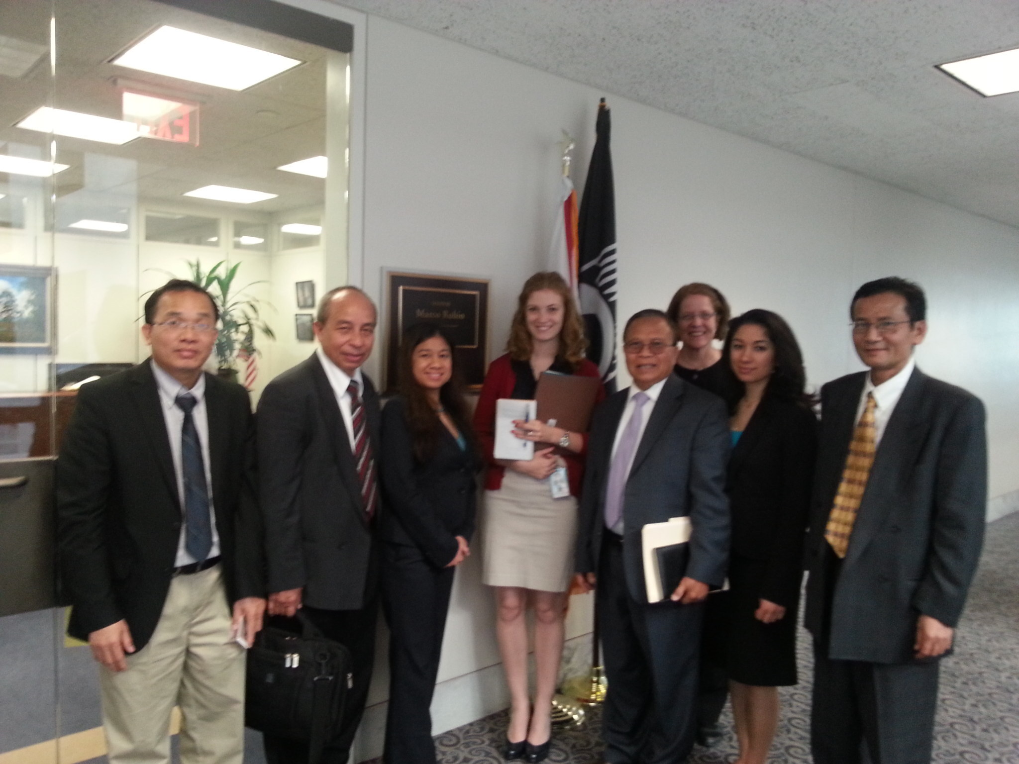 A group of men and women in business clothes pose in a congressional office lobby with flags behind them