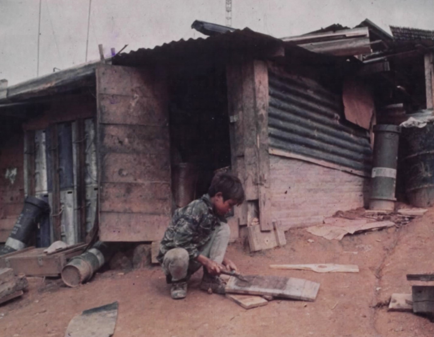 A boy crouching outside a small house of timber and corrugated iron