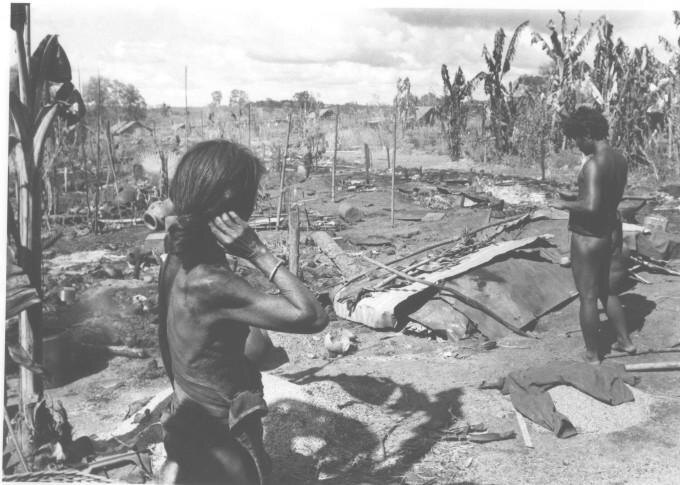 Two people standing in the ashes of burned houses, bodies on the ground before them