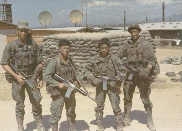 Four soldiers with rifles standing in front of sandbagged positions at a camp