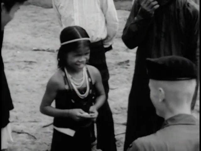 A smiling highland girl with a beaded headband and necklaces stands among soldiers