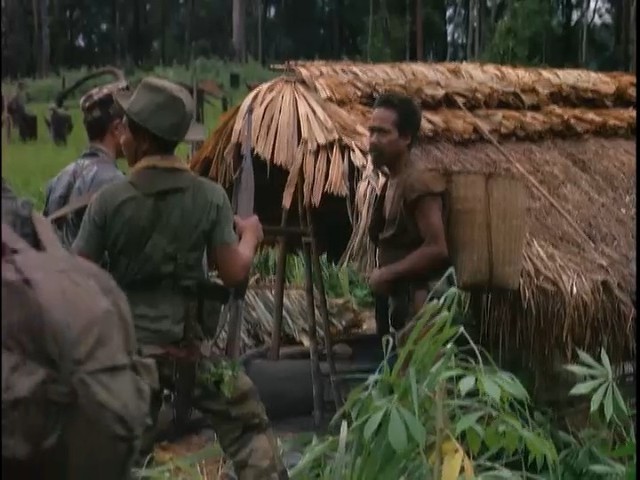 A highland man in a loincloth stands by a thatched house while a soldier in a helmet questions him