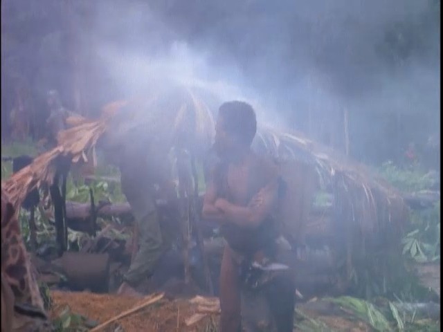 A highland man stands beside a smoking thatched hut, with soldiers moving behind him