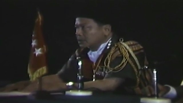 A highland delegate in a woven jacket with braided cords sits at a table with microphones, a flag behind him