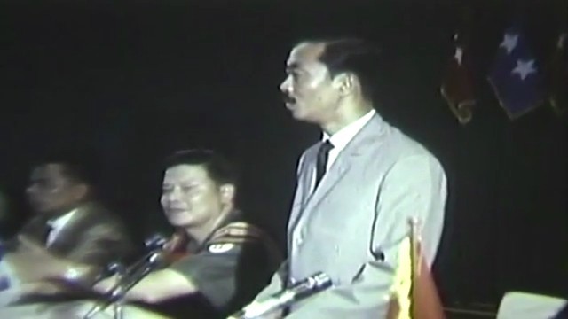 A man in a gray suit speaks at a microphone beside delegates in woven highland jackets, with flags behind them