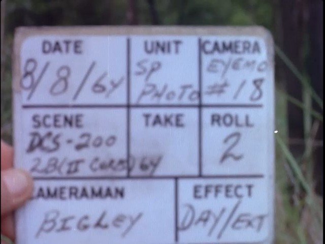A hand holds up a film slate marked 8/8/64, SP Photo, Eyemo 18, II Corps, cameraman Bigley