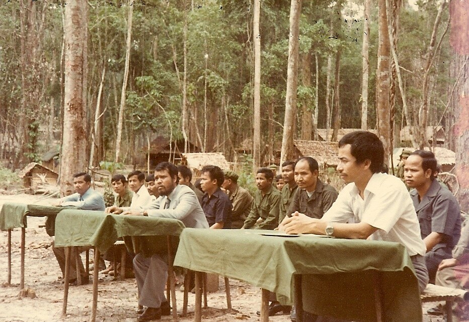 Men in plain shirts seated at tables covered with green cloth in a forest clearing, with huts behind