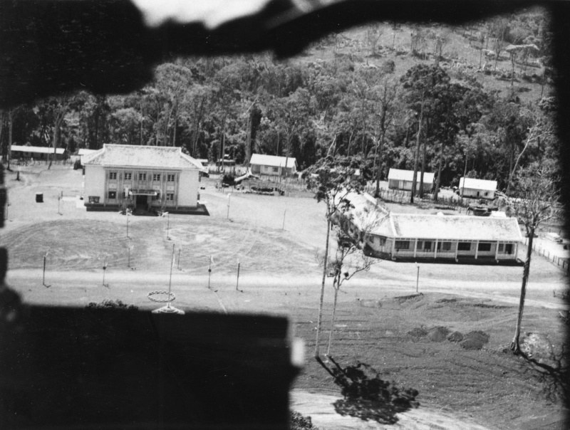 A black and white aerial photograph of a few buildings in a clearing surrounded by forest