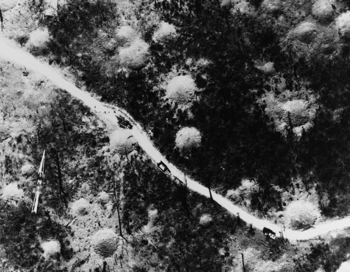 An aerial photograph of a winding road through forest with bomb craters and small trucks