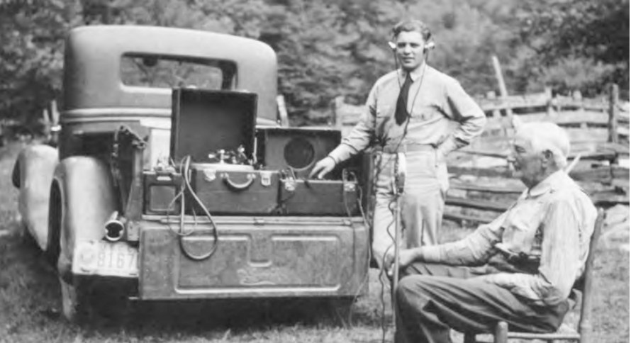 A young man wearing headphones stands beside recording equipment in the back of a car, while an old man sits on a chair in front of a microphone