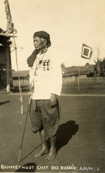 An elderly man in a turban and white jacket with medals, standing barefoot with a spear