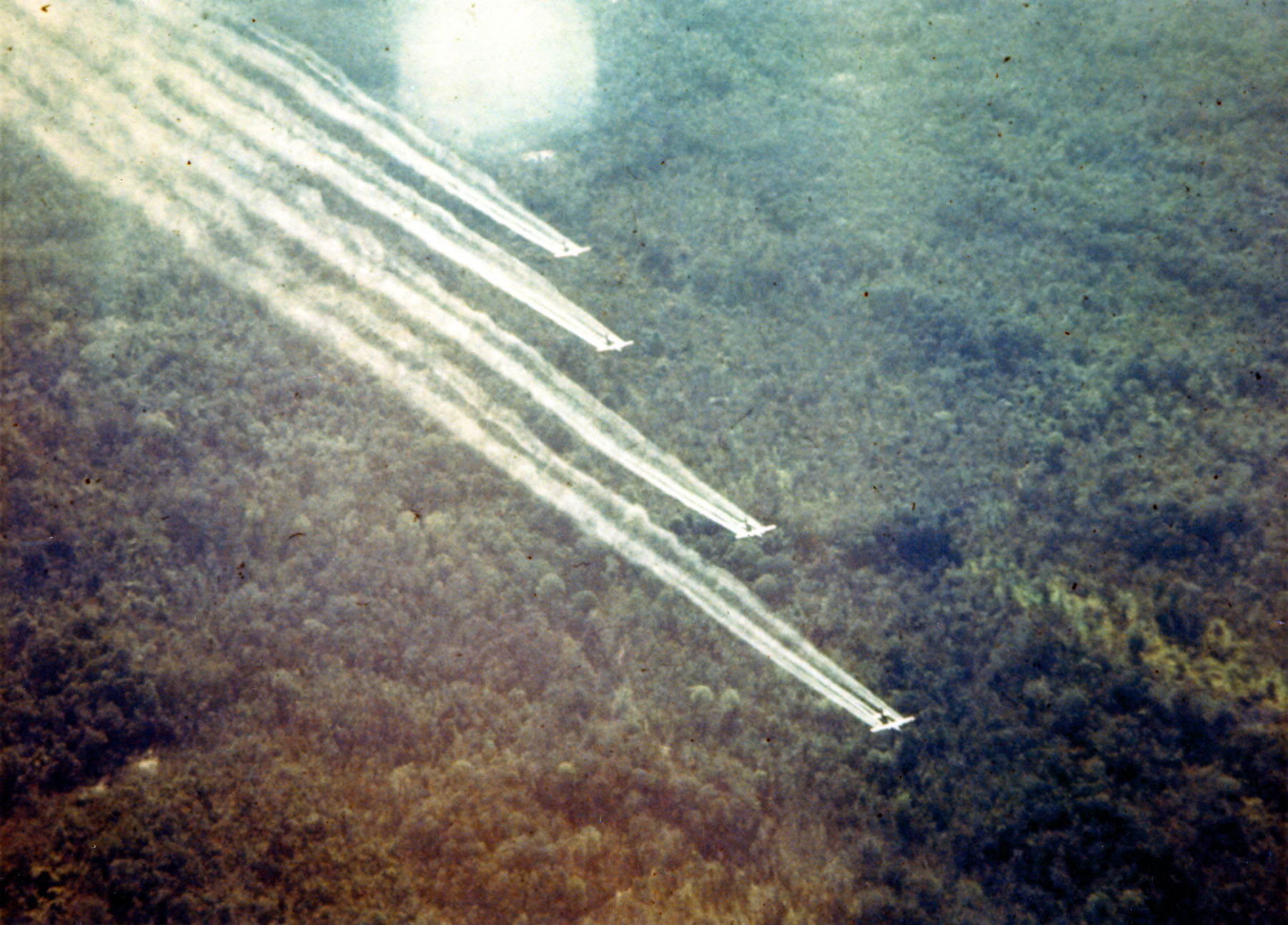 Four aircraft flying in formation low over forest, trailing spray