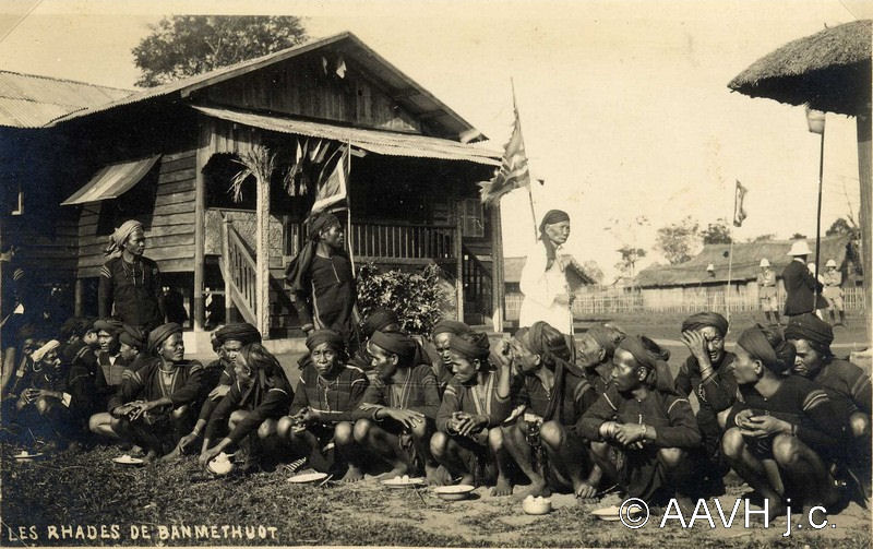 A large group of Rhade men and women seated in front of a stilt house