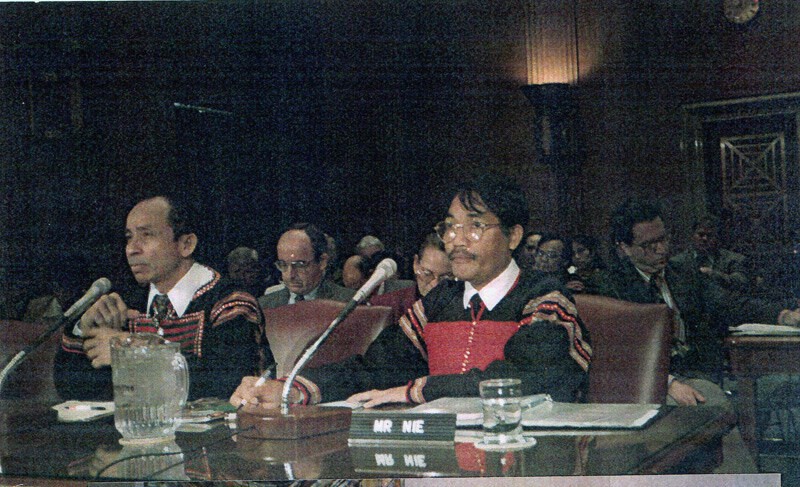 Two highland men in woven jackets seated at a witness table with microphones