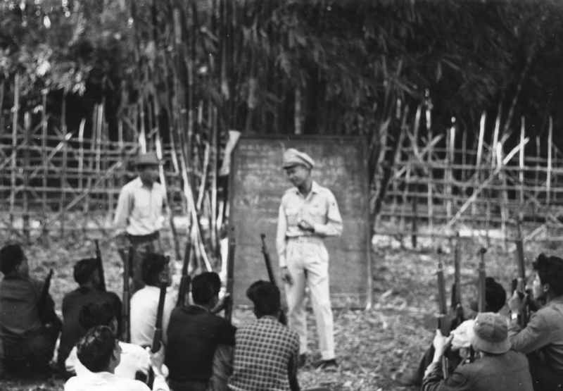 An American in a white shirt stands before armed villagers seated on the ground beside a bamboo fence