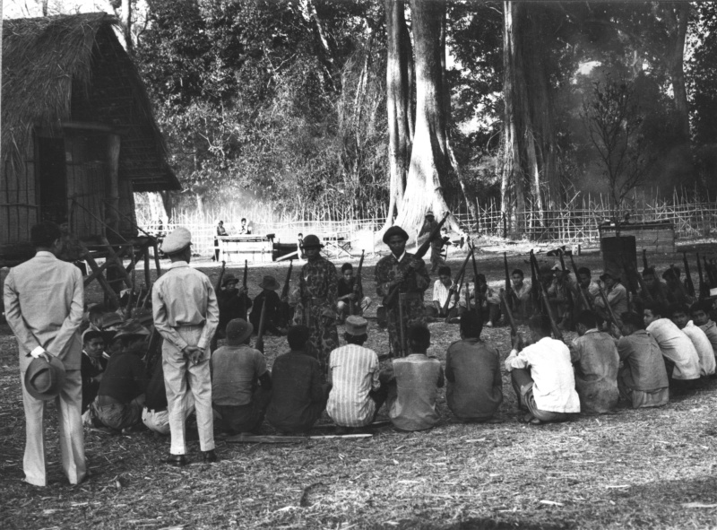 Armed men seated in a circle in a village clearing with two soldiers standing among them