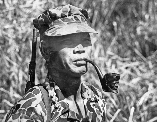 Y Bham Enuol in camouflage and a soft cap, a pipe in his mouth, a rifle slung over his shoulder