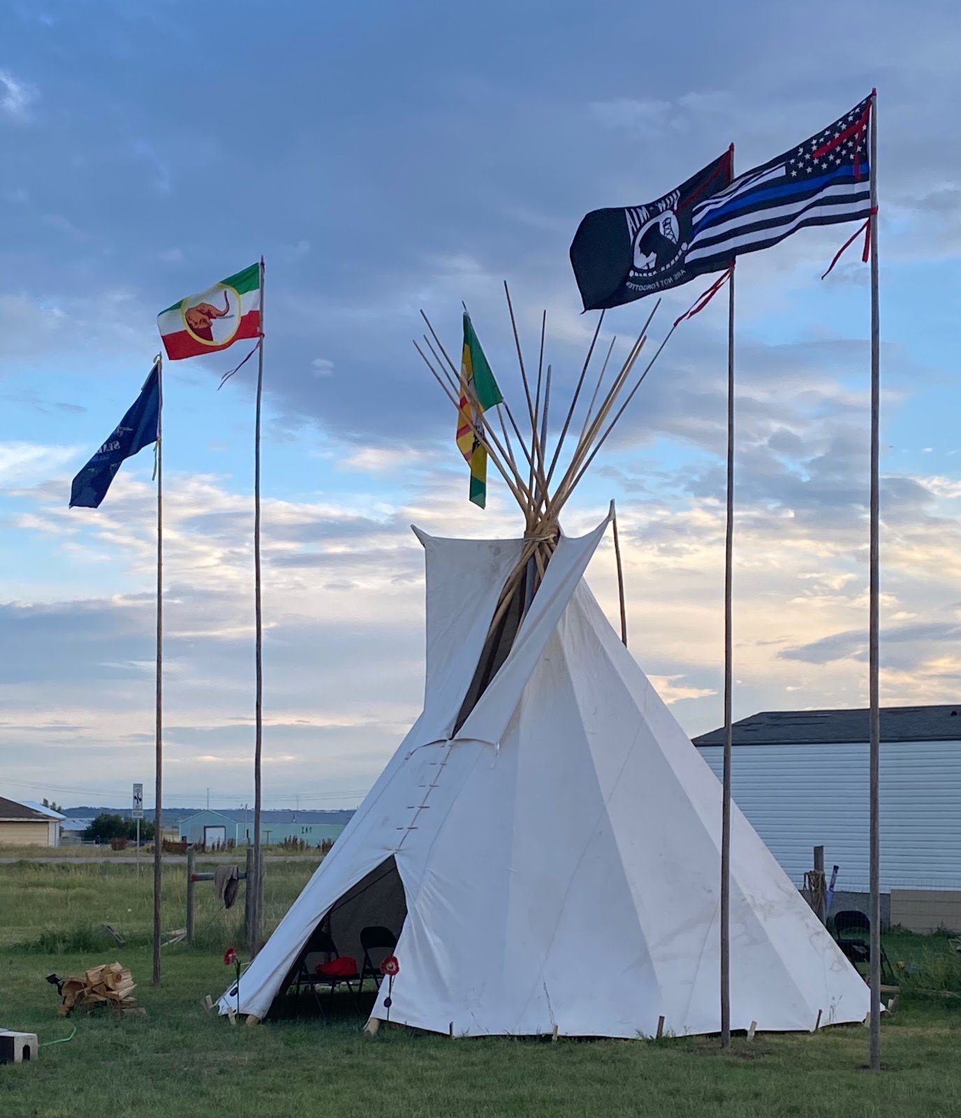A white teepee pitched on grass at dusk with several flags flying on poles around it, including the POW/MIA flag