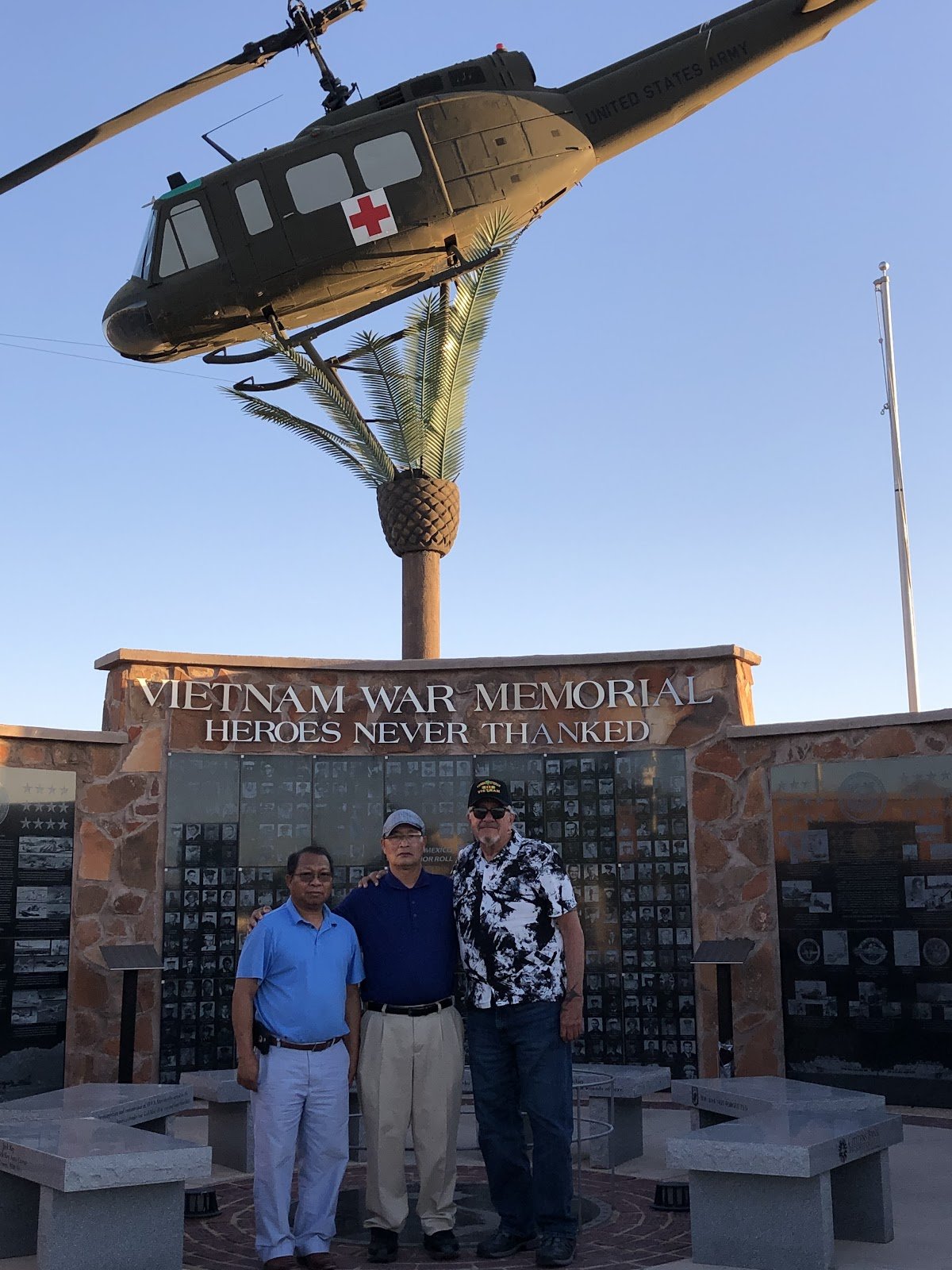 Three men standing in front of the Vietnam War Memorial wall in Las Cruces, beneath a US Army Huey helicopter mounted above it, under the words Vietnam War Memorial: Heroes Never Thanked