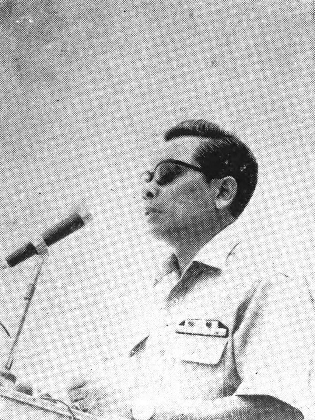 Paul Nur in glasses and a light short-sleeved shirt, speaking into a microphone at a lectern