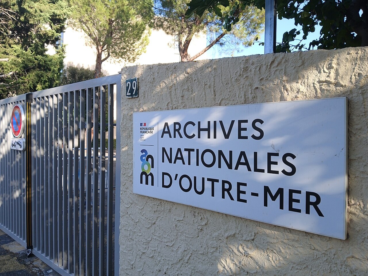 A metal gate in a garden wall beside a sign reading Archives nationales d’outre-mer