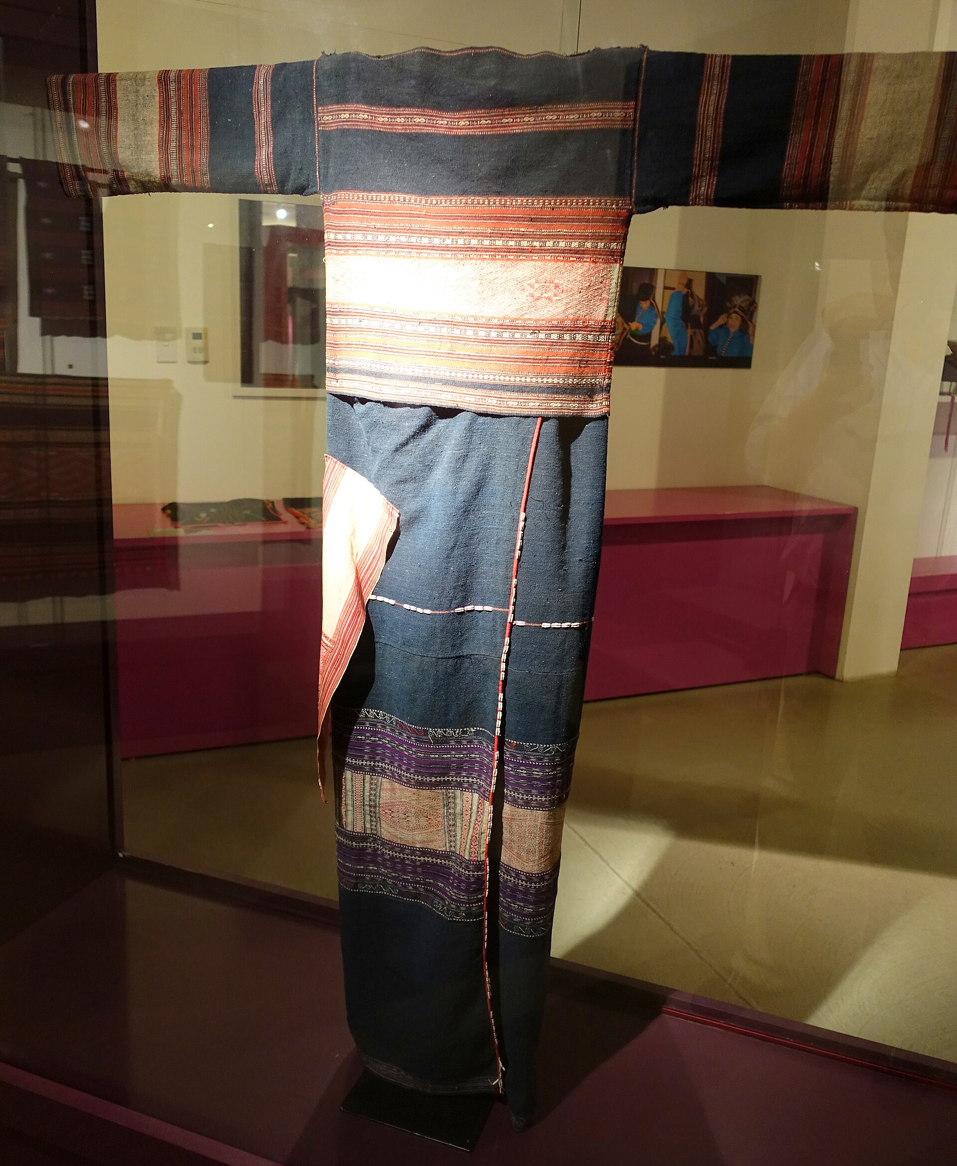 A Bahnar shirt and skirt in dark cotton with woven bands, displayed in a museum case