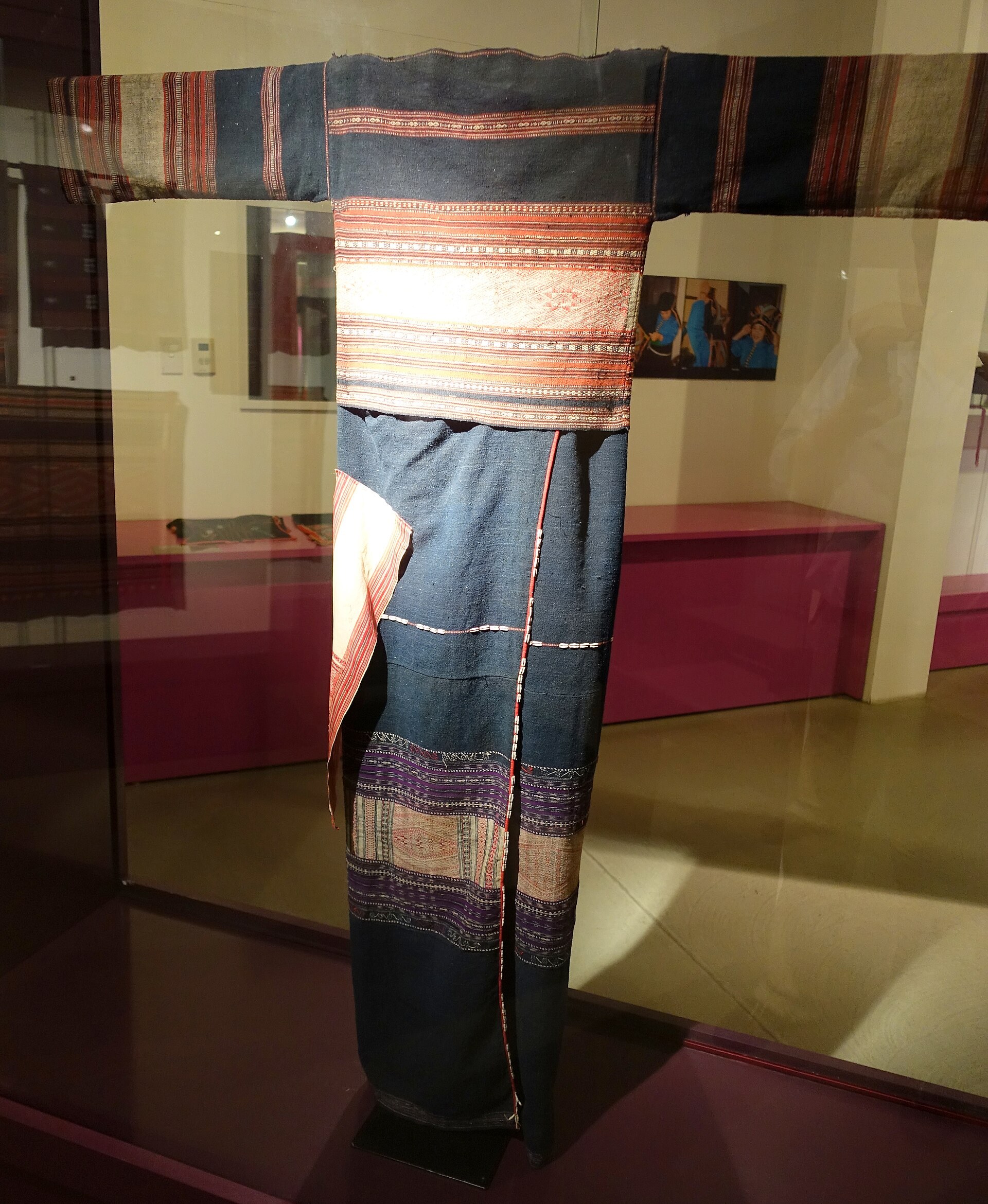 A Bahnar woman’s dark costume with red patterned bands and a skirt hung with seeds, on a museum figure