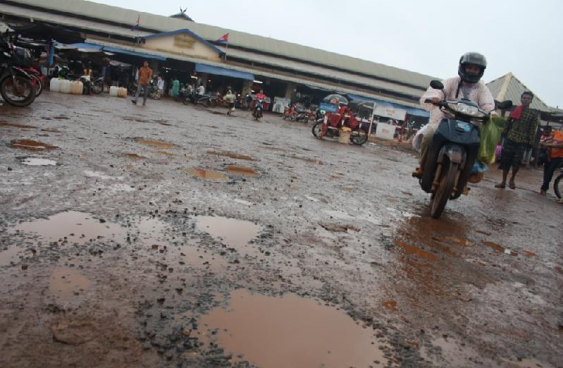 A muddy square in front of a market hall, with motorbikes passing