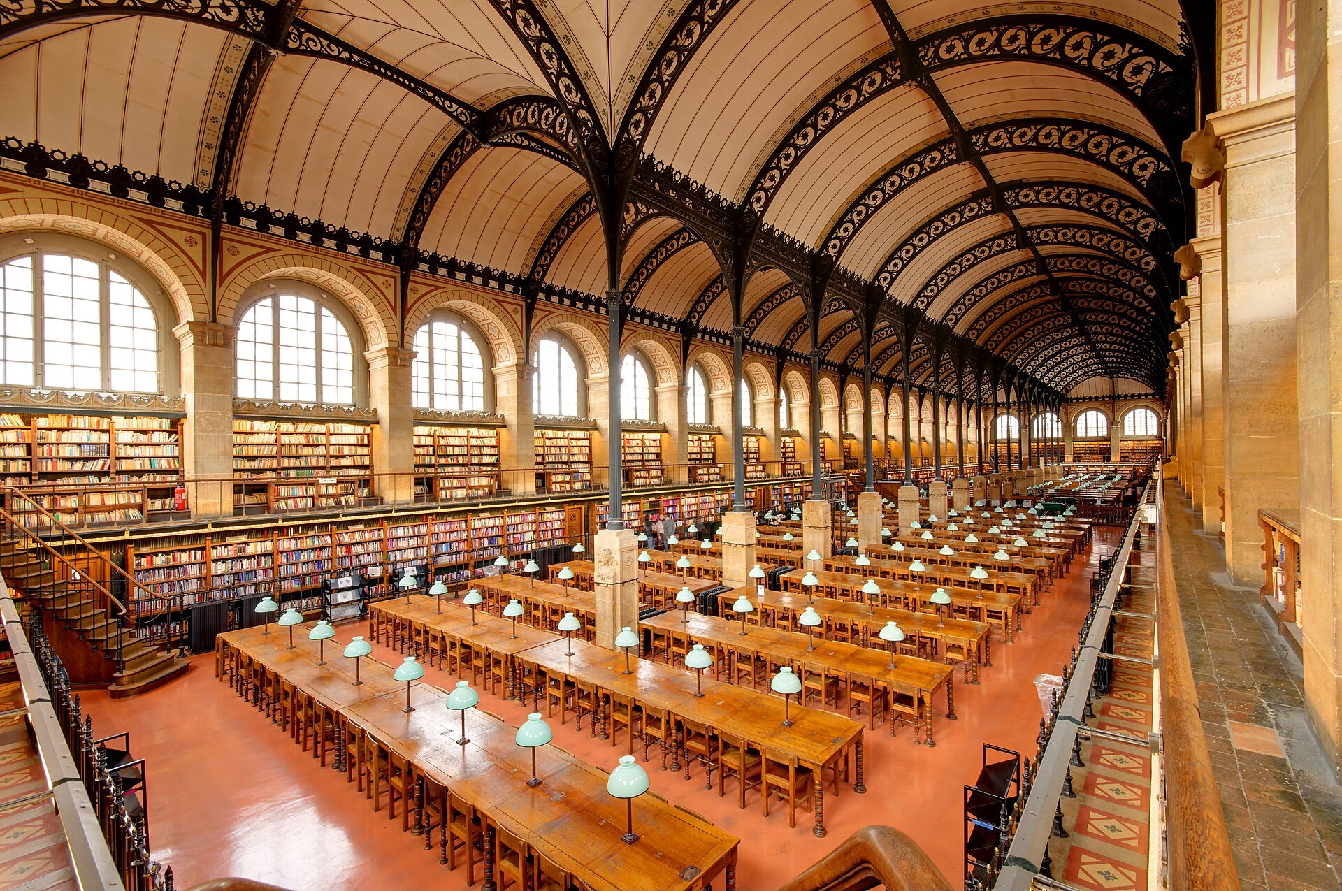 A long reading room with an iron-vaulted ceiling, rows of desks with green lamps and bookshelves along the walls