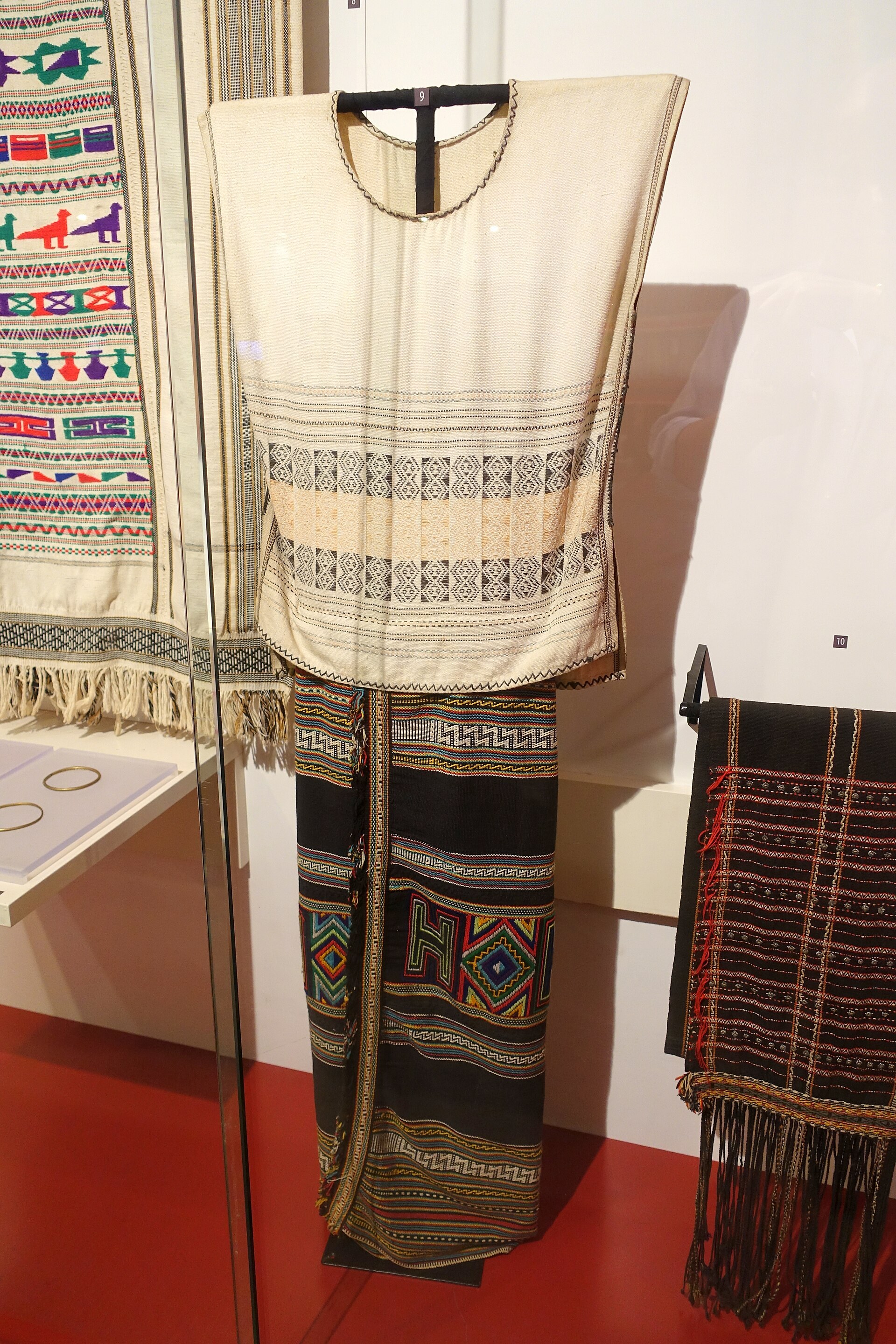 A bride’s dark costume with red and white bands, from Bao Lam in Lam Dong, on a museum figure
