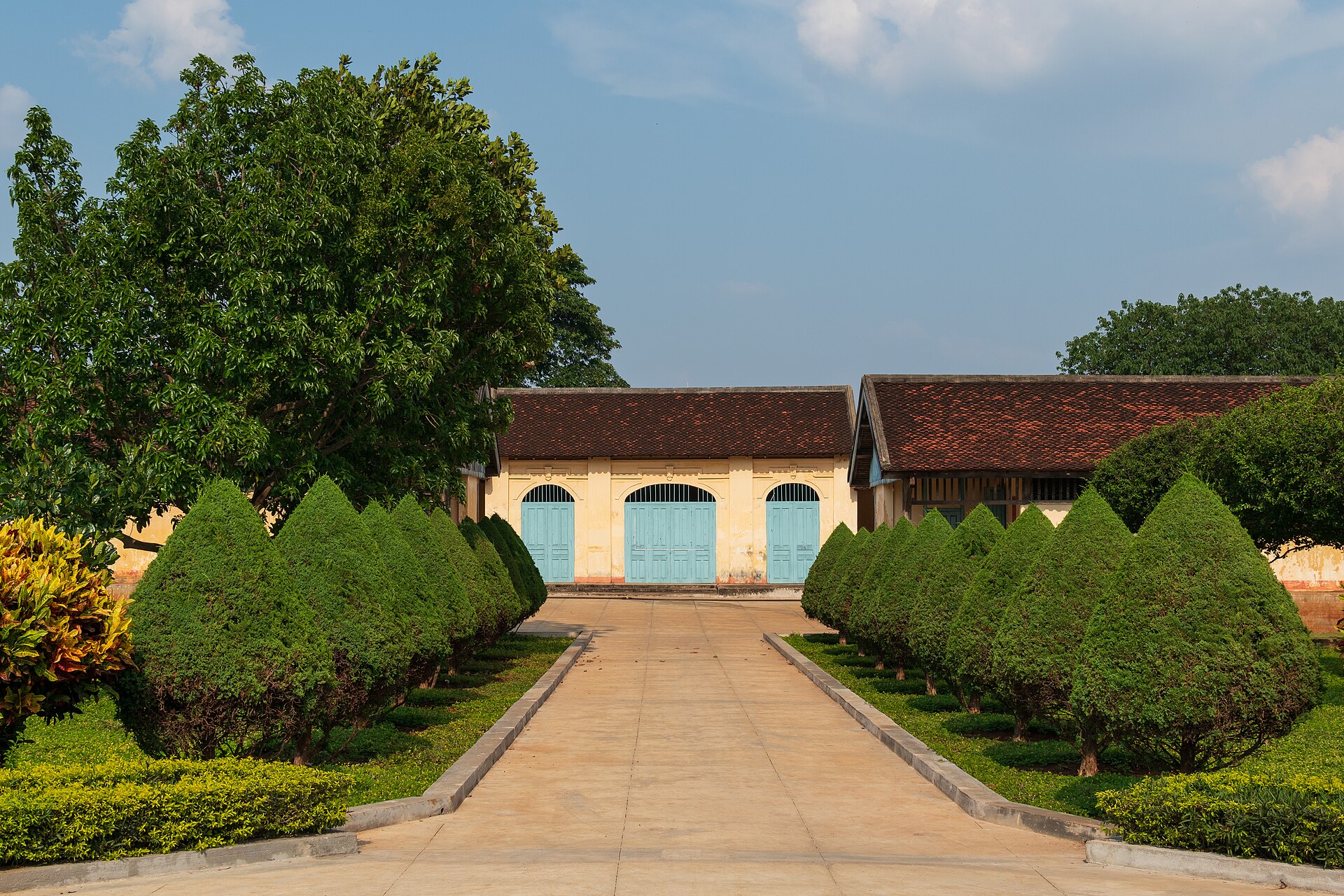 A low yellow colonial building with blue doors at the end of a path lined with clipped hedges