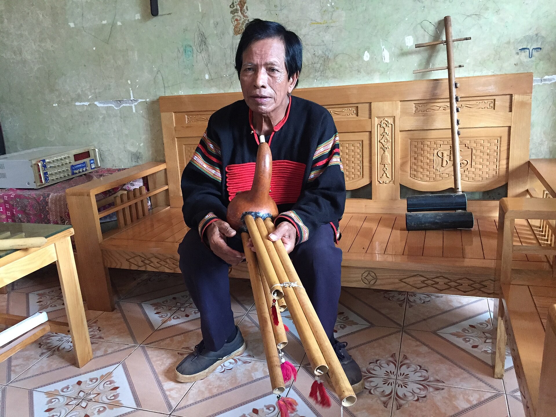 A Rhade man seated indoors playing a gourd mouth organ with long bamboo pipes