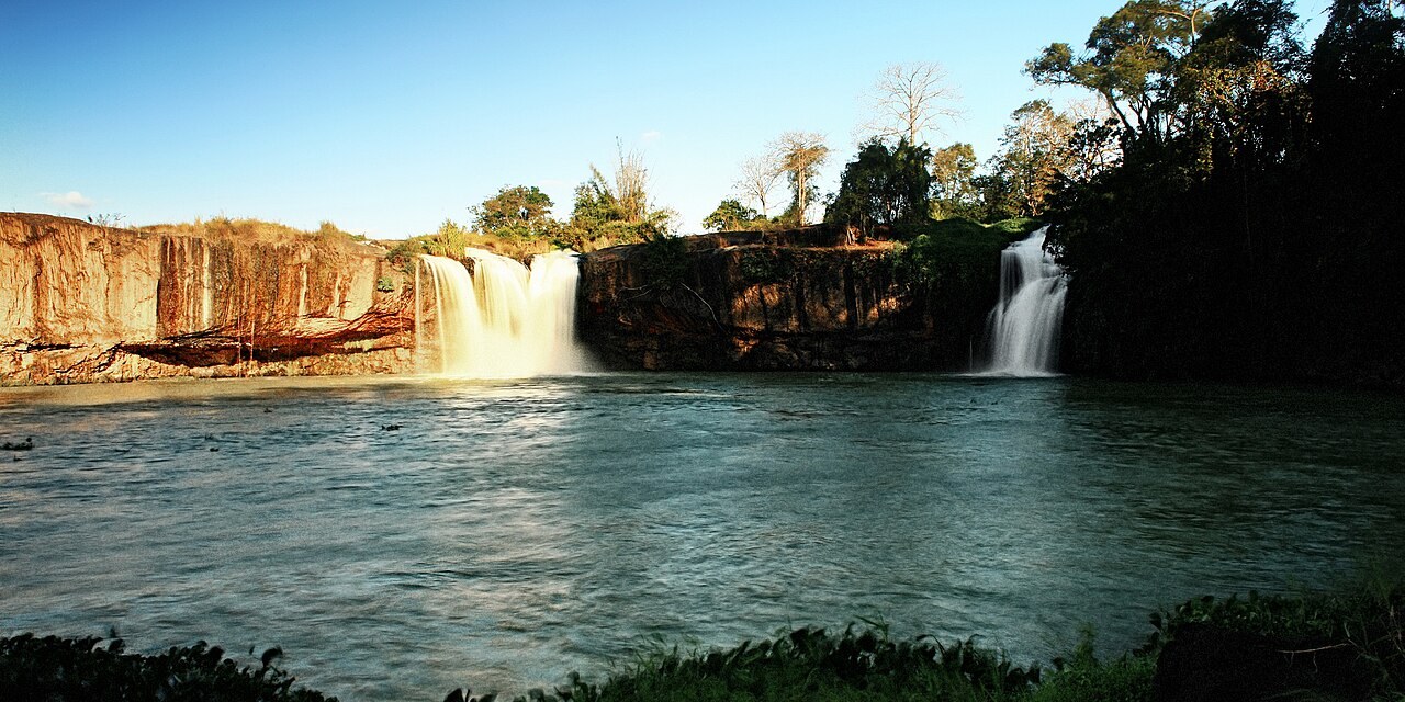 A wide waterfall pouring over a rock ledge into a pool, with forest on either side