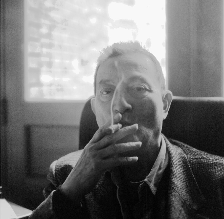 A black and white portrait of an older man smoking, seated at a desk