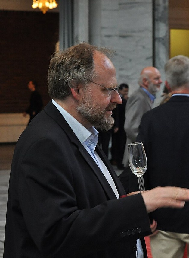 A man with glasses and a dark jacket speaks at a reception, holding a glass
