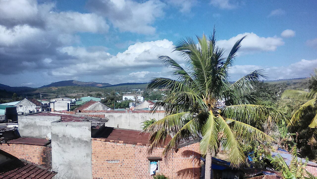 Tiled roofs of a village among palm trees under a cloudy sky, with hills beyond