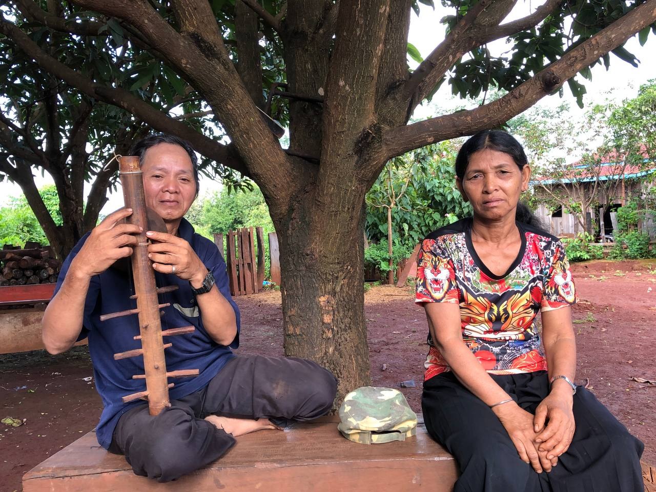 An older Jarai man sits under a tree playing a long bamboo string instrument, with his wife beside him