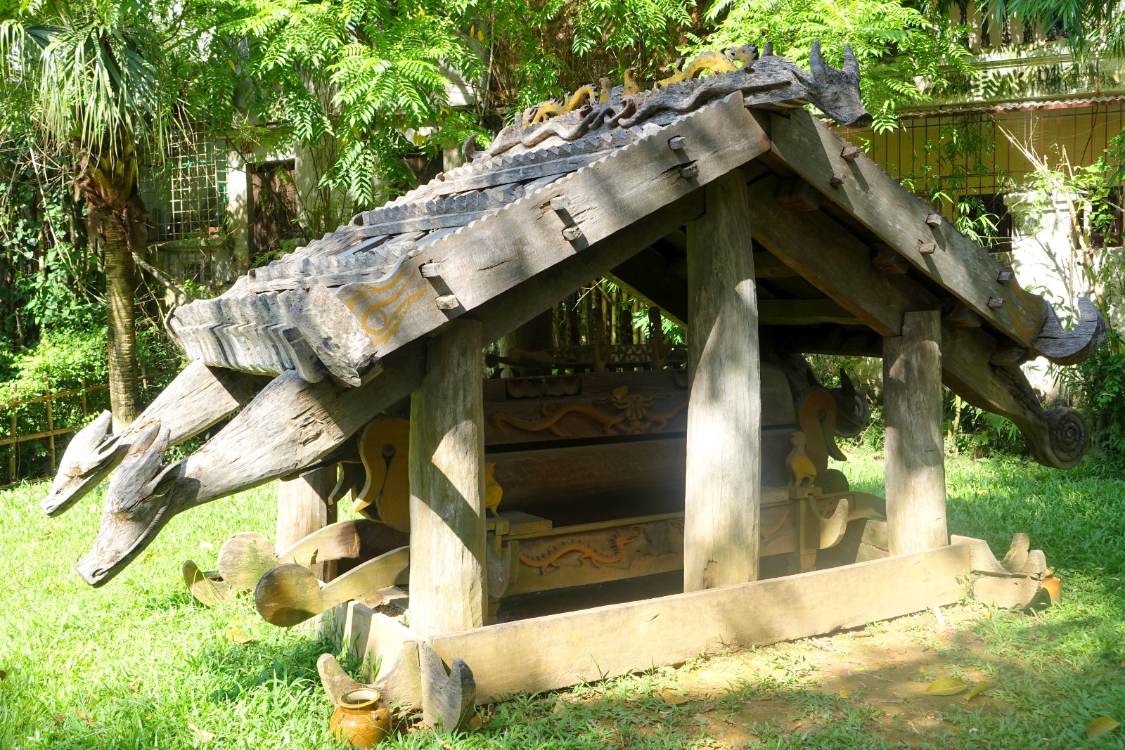 A carved wooden tomb house with animal figures on the roof, in a museum garden