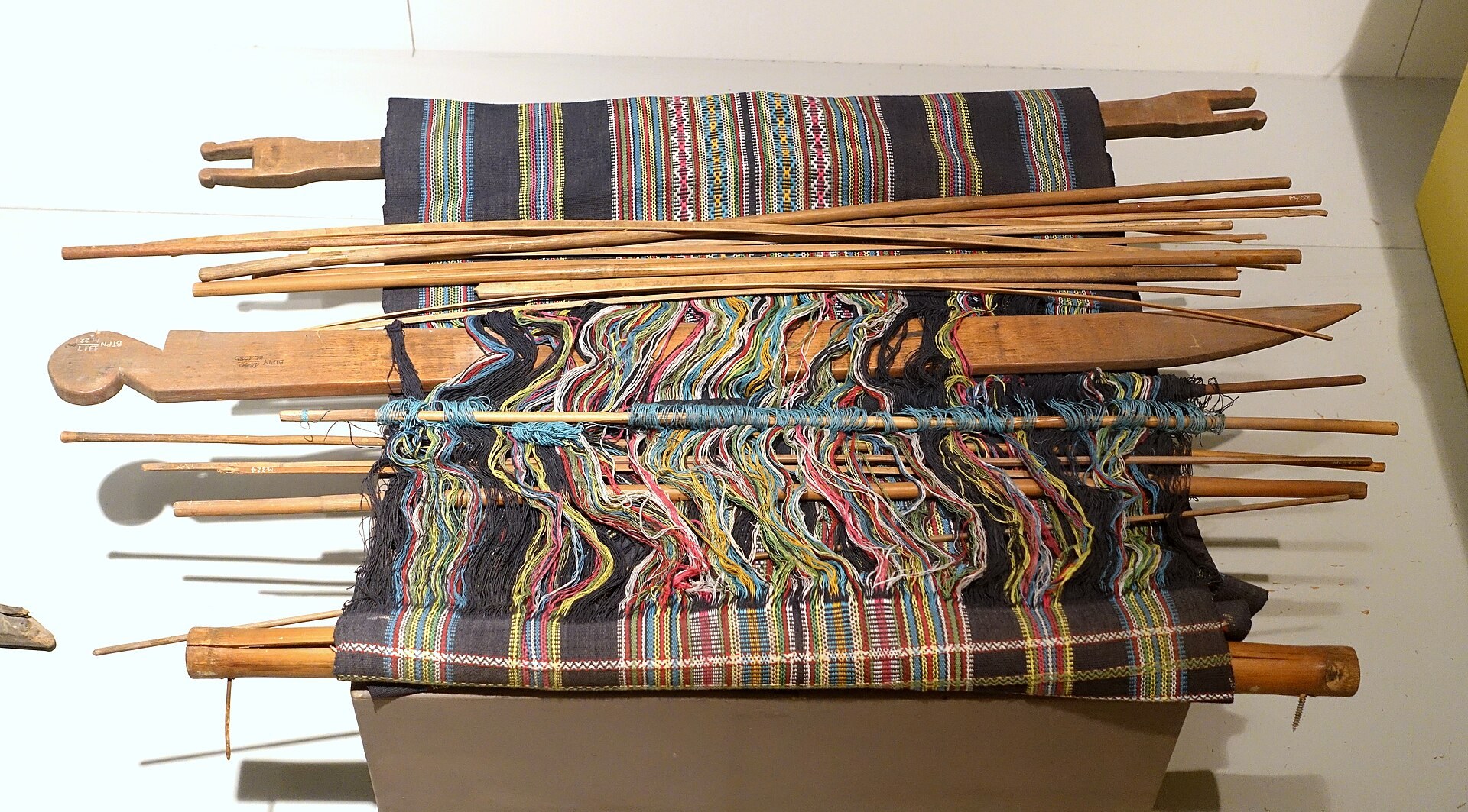 A backstrap loom with a partly woven striped cloth, the rods, sword and beams laid out in a museum case