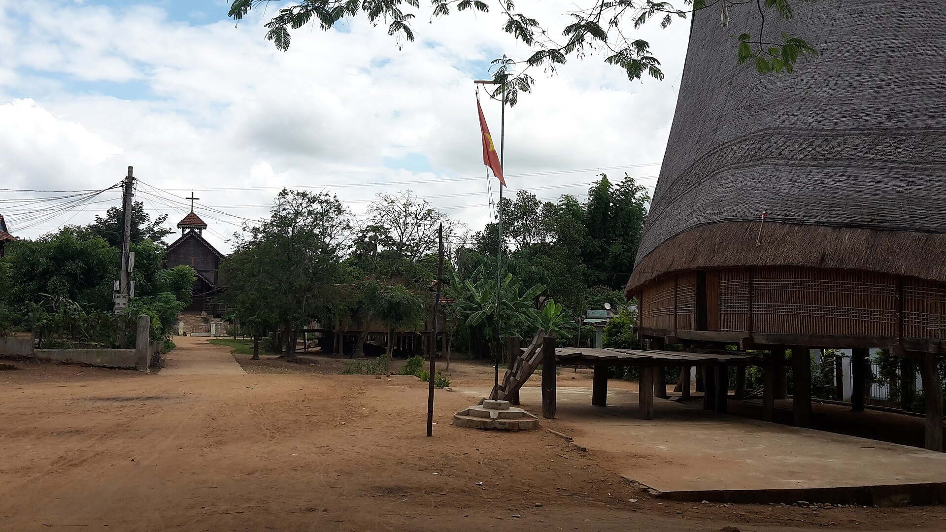 A Bahnar village square with a tall thatched communal house on stilts among trees