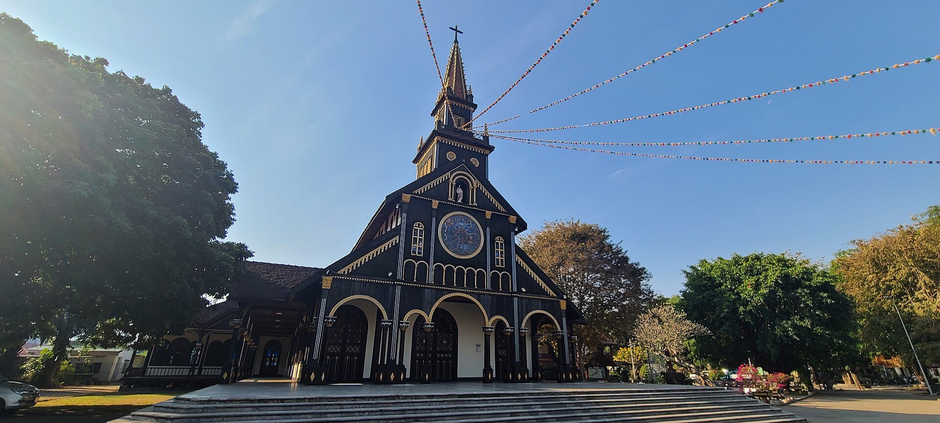 A dark wooden church with a tall central spire, a rose window and a wide flight of steps under strings of flags