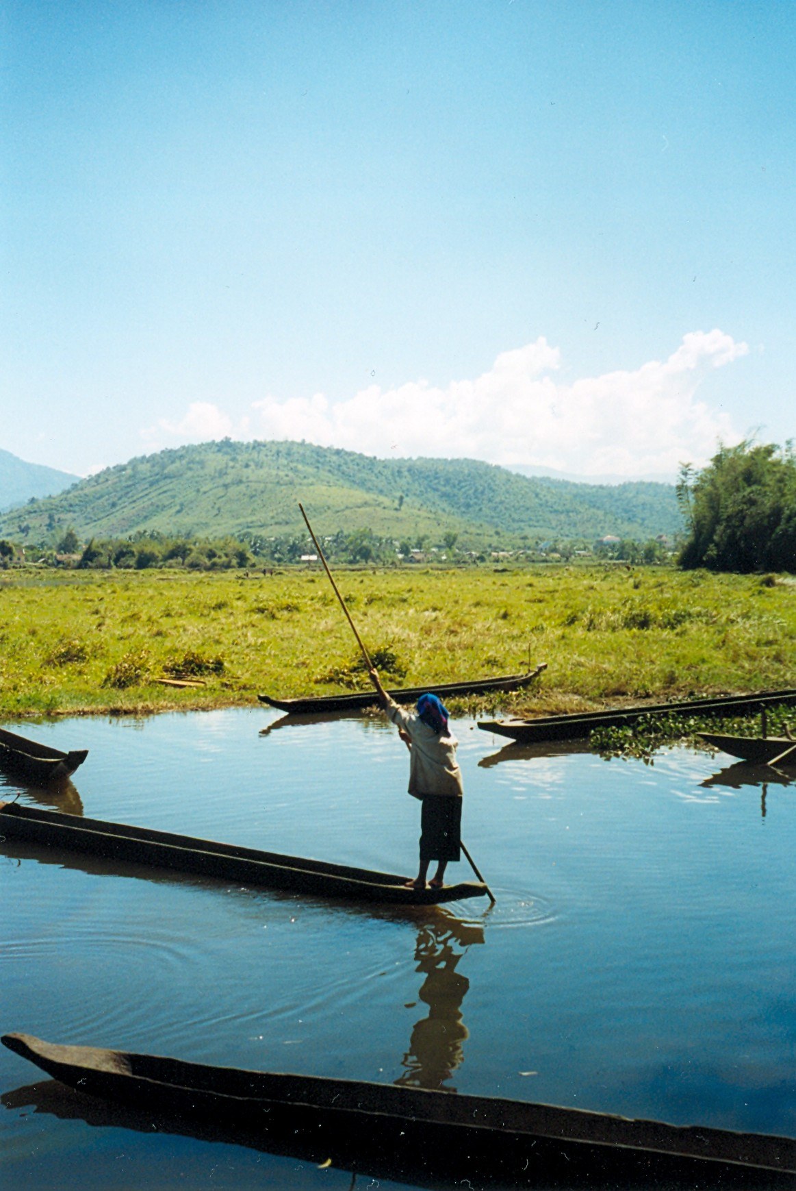 A man standing in a dugout canoe on a lake covered in water plants, with hills behind
