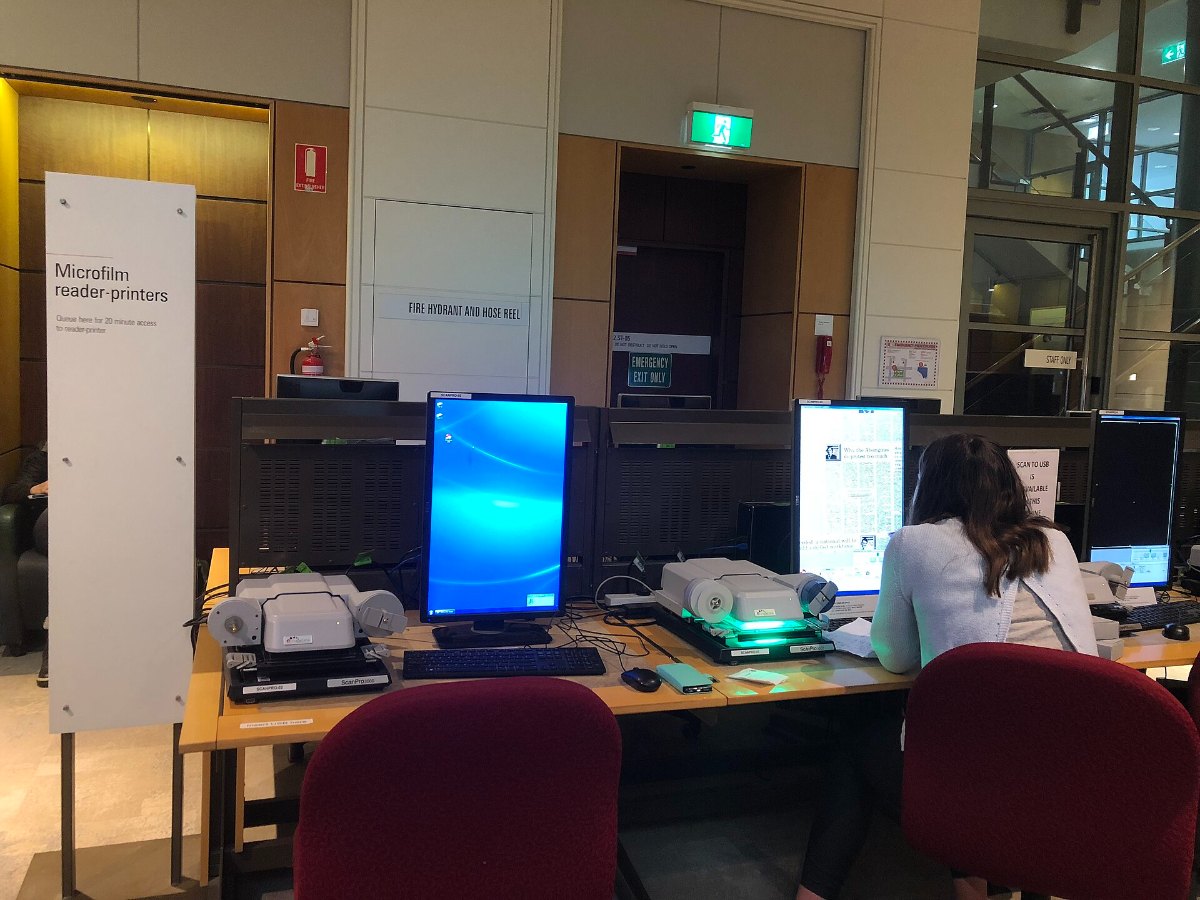 A row of microfilm reader-printers on desks in a library, with a reader seated at one of them
