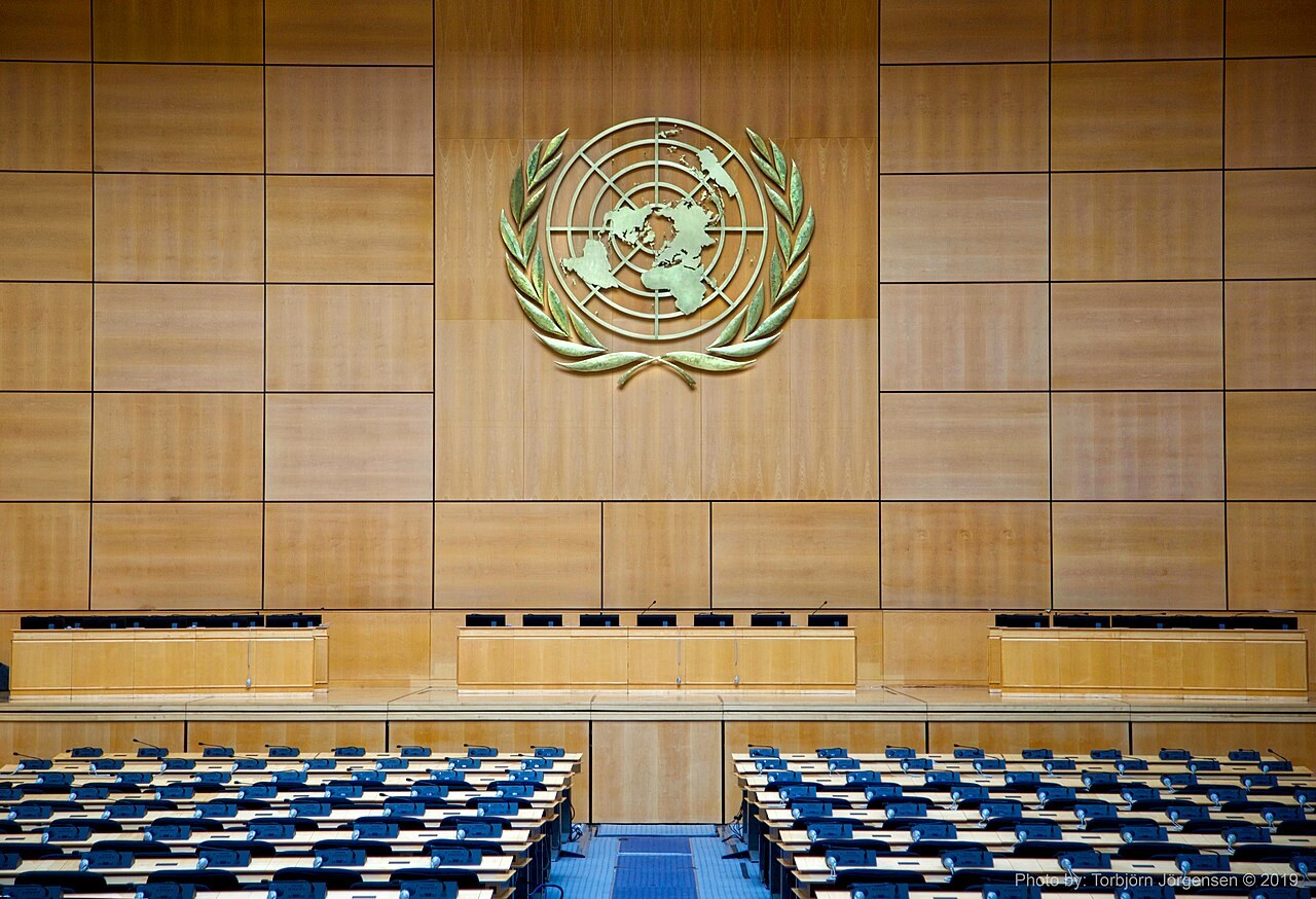 A large assembly hall with wood-paneled walls, the UN emblem above the podium and blue rows of seats