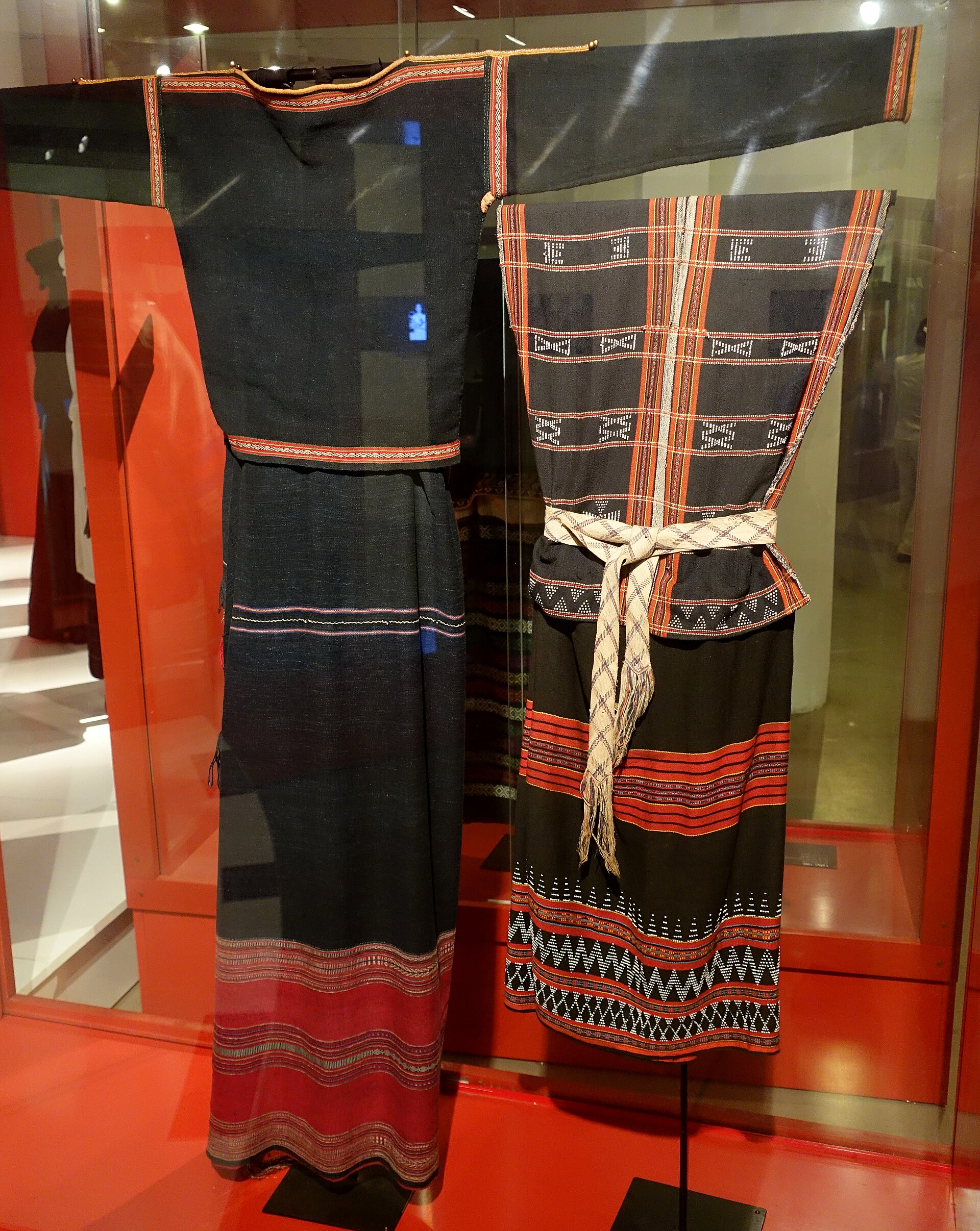 A dark handwoven costume with red and white bands in a museum display case