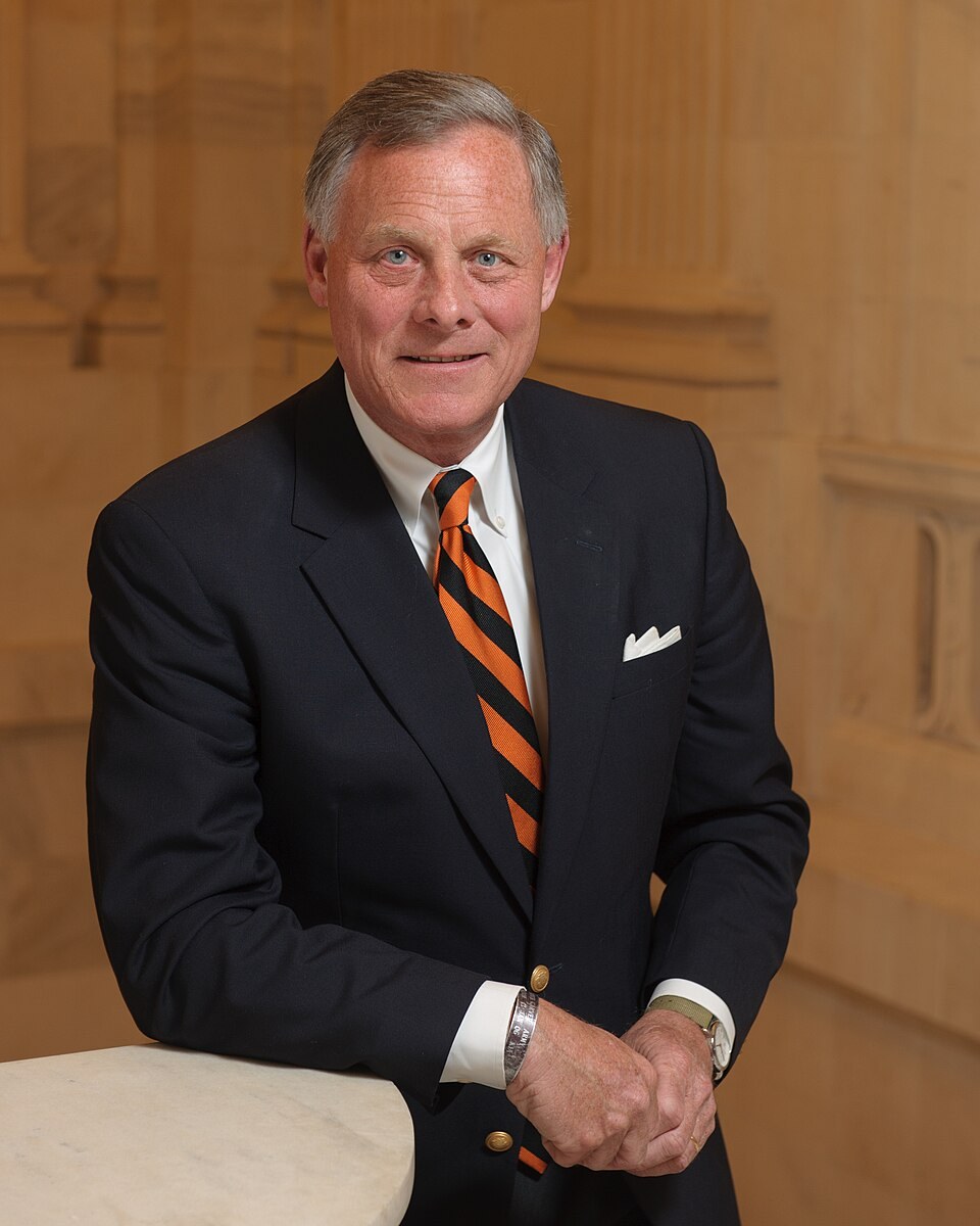 An official portrait of a gray-haired man in a dark suit and striped tie