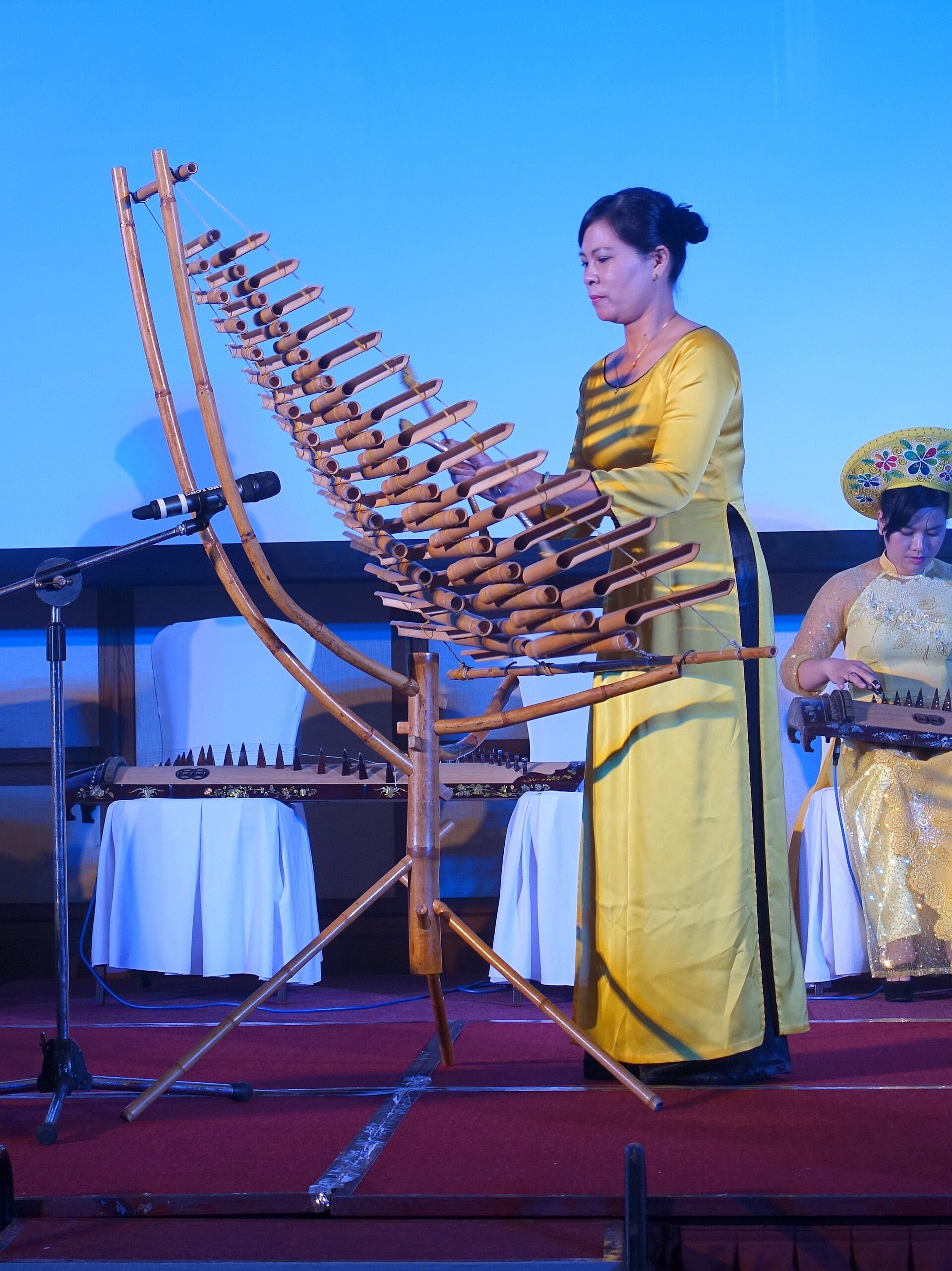 A performer striking a large bamboo xylophone with two sticks, the tubes hung in a curved frame