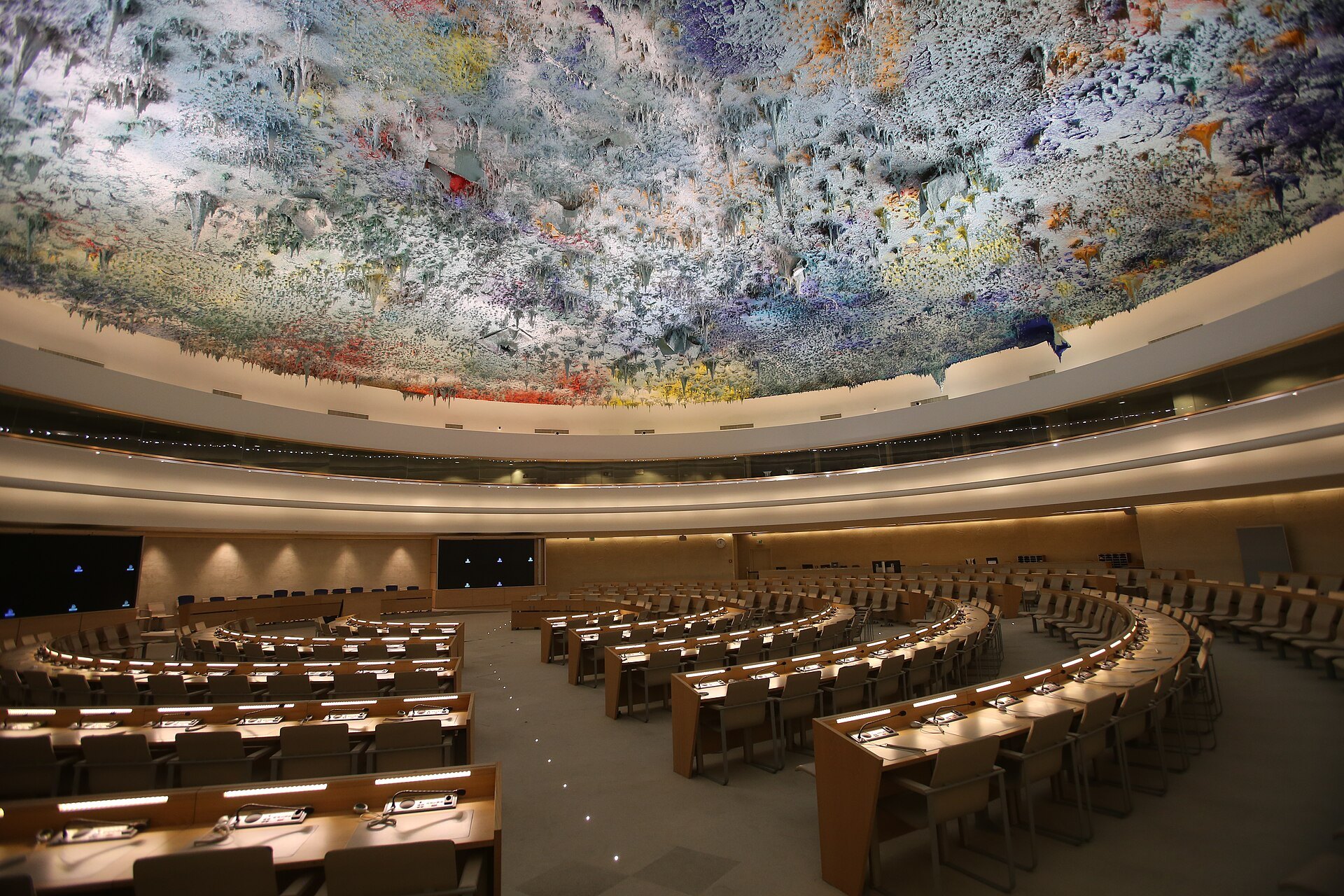 A large round conference chamber with curved rows of desks under a sculpted, brightly colored ceiling