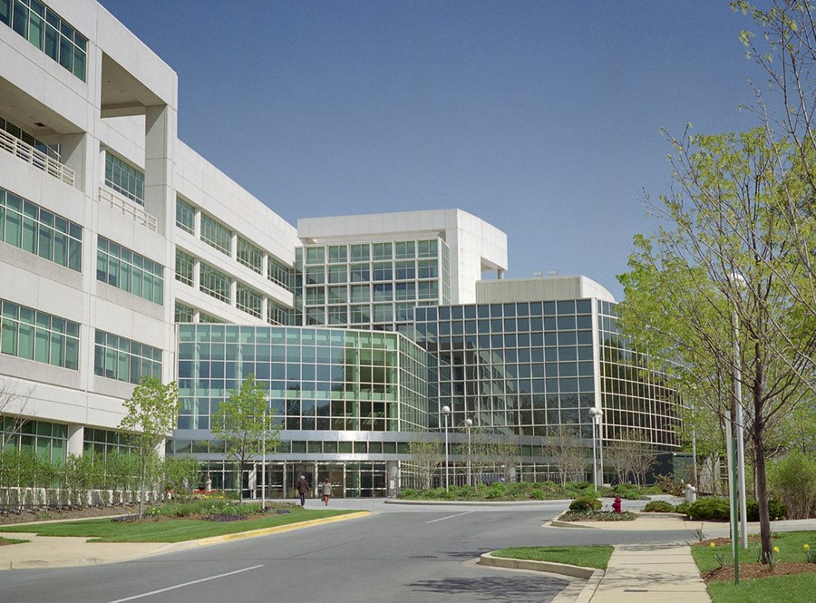 A large modern building of glass and pale stone beside a road and trees