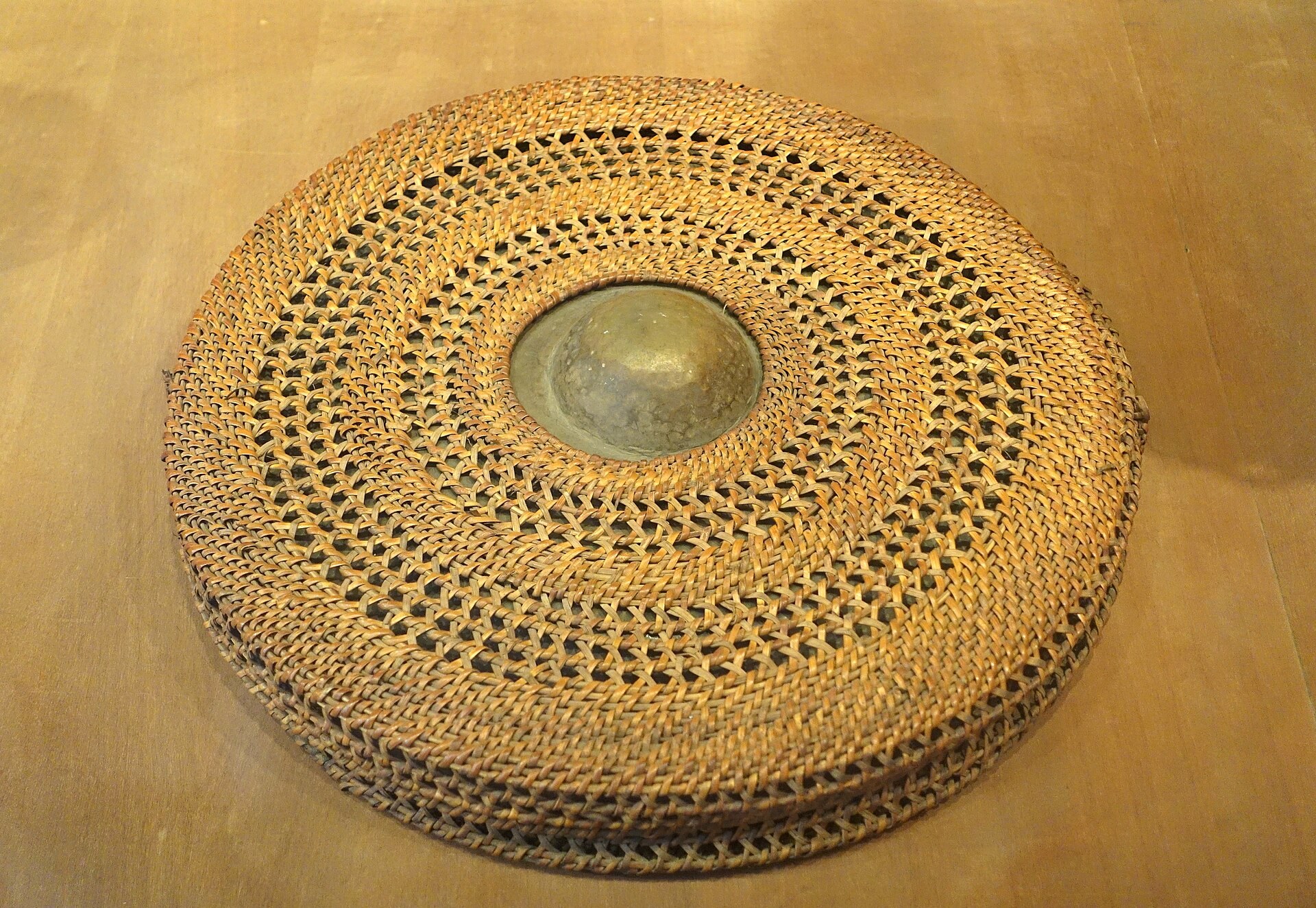A bossed bronze gong beside its woven rattan carrying case, on a museum stand